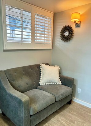 Un sofá tapizado en gris con una almohada decorativa blanca, frente a una pared con una lámpara de pared y un reloj decorativo.