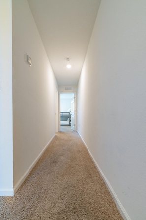 A narrow hallway with carpeted floors and white walls, leading to a room with a bunk bed. - Modern 3BR Townhome | 30-Day Minimum (Jacksonville)