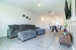A modern living room with a grey sectional sofa, a matching ottoman, a dining area with a table and chairs, and a small side table with a vase. - Modern 3BR Townhome | 30-Day Minimum (Jacksonville)