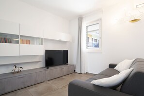 Ein modernes Wohnzimmer mit einem Flachbildfernseher, integrierten Regalen mit Büchern, einem grauen Sofa und Blick ins Freie.