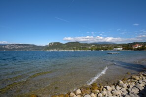 A clear lake with a rocky shoreline, boats, and a distant cityscape with mountains. - Apartment 'Dama' with Private Terrace, Wi-Fi and Air Conditioning (Bardolino)