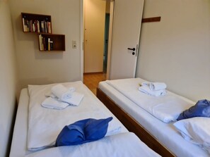 A hotel room with two single beds, each with a neatly folded towel and a pillow. There is a bookshelf with books and a door leading to another room. - 12 / Waldstrasse (Wyk auf Foehr)