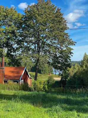 A rural landscape with a red-roofed building, a large tree, and a body of water in the distance. - Cottage by Lake Ralången with alpacas and horses (Aneby)