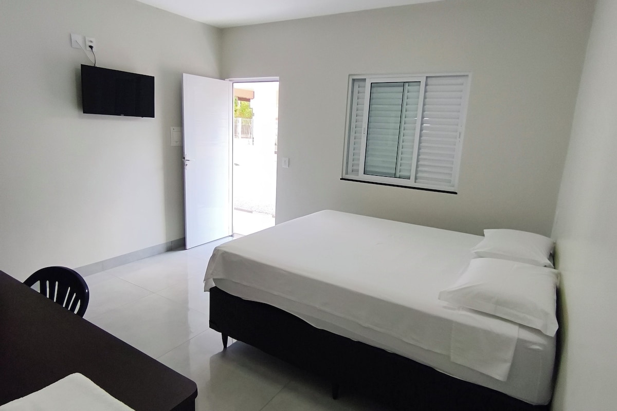 Een hotelkamer met een bed, een bureau, een stoel, een aan de muur bevestigde televisie en een raam met jaloezieën.
