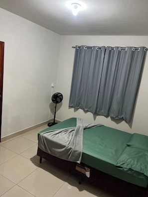 Habitación con una cama individual, colchón verde, manta gris, ventilador de pie y cortinas grises.