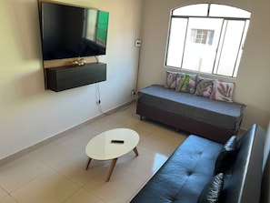 Una sala de estar con una televisión de pantalla plana montada en la pared, un sofá, una pequeña mesa redonda y una ventana con vistas a edificios.