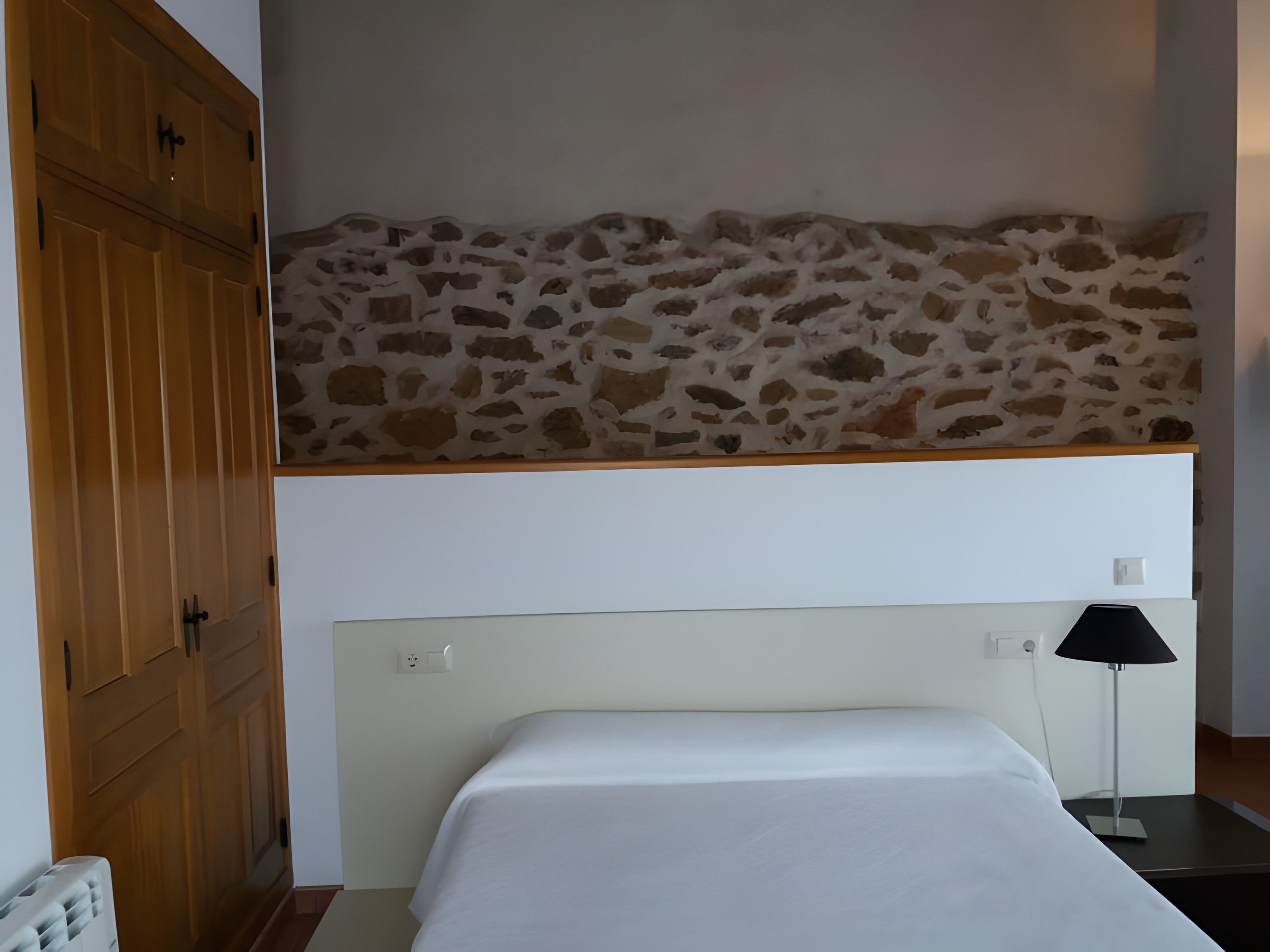 Een slaapkamer met een stenen muur, een bed, een nachtkastje en een houten garderobe.