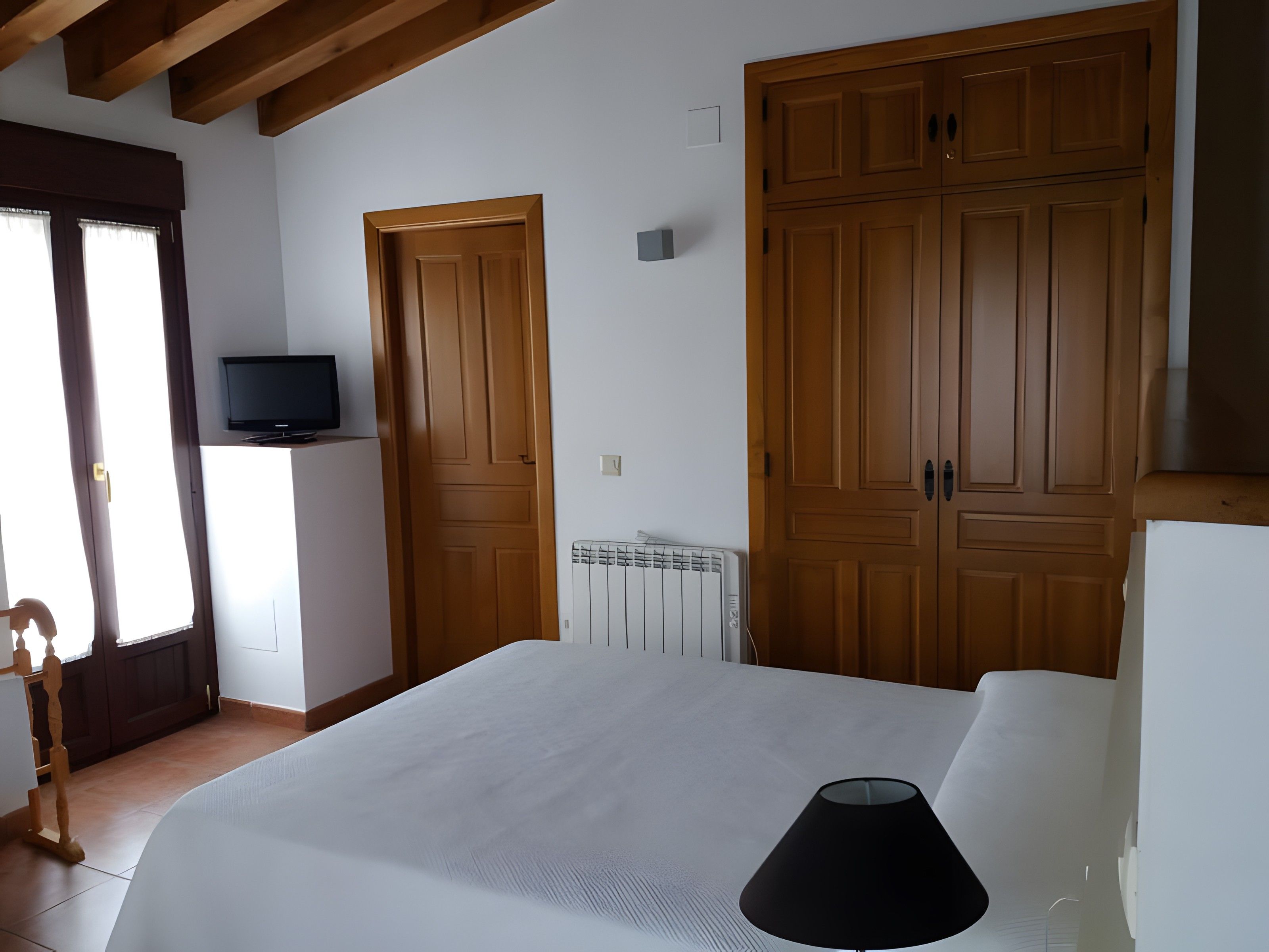 A bedroom with a bed, a lamp, a television on a shelf, wooden wardrobes, and a radiator.