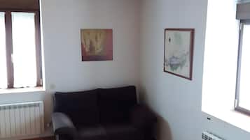 Ein Wohnzimmer mit Holzboden, einem schwarzen Sofa, einem Glastisch und zwei mit Vorhängen versehenen Fenstern.