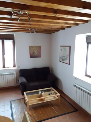 Een woonkamer met een houten plafond, een zwarte bank, een glazen salontafel en twee ramen met gordijnen.