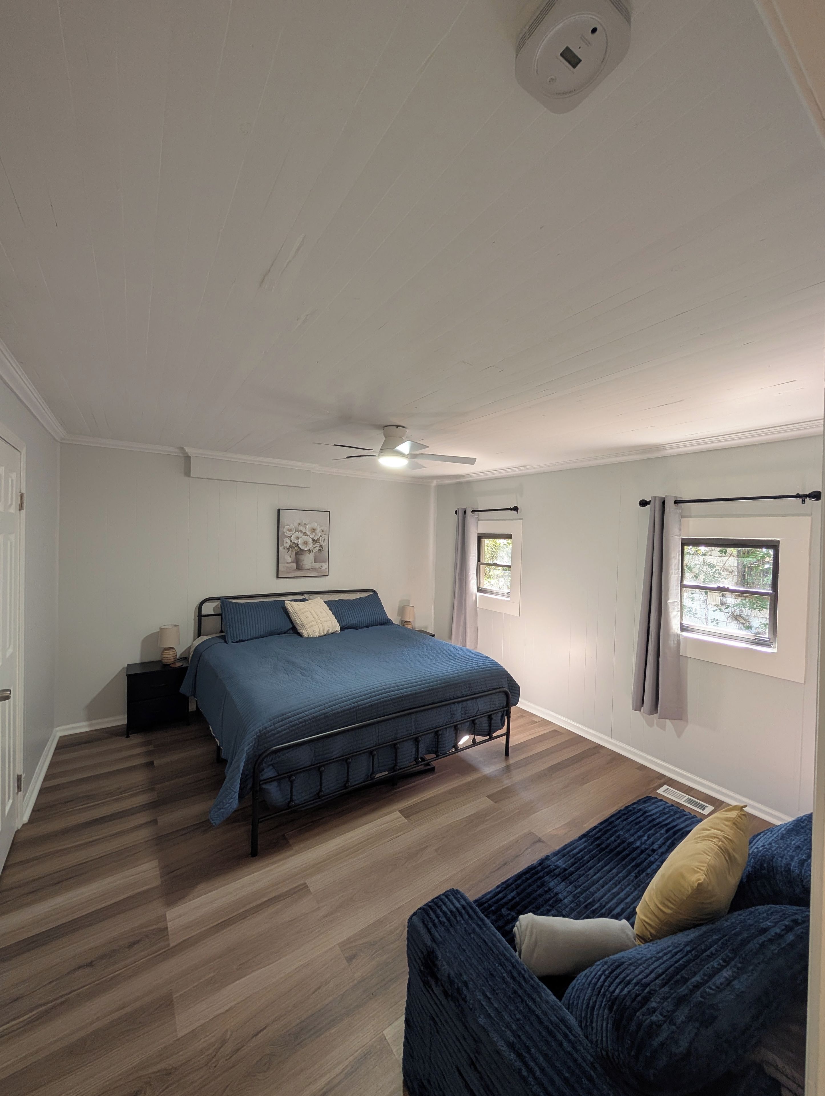 Un dormitorio con cama, mesitas de noche, ventilador de techo, ventana con cortinas y vistas al exterior.