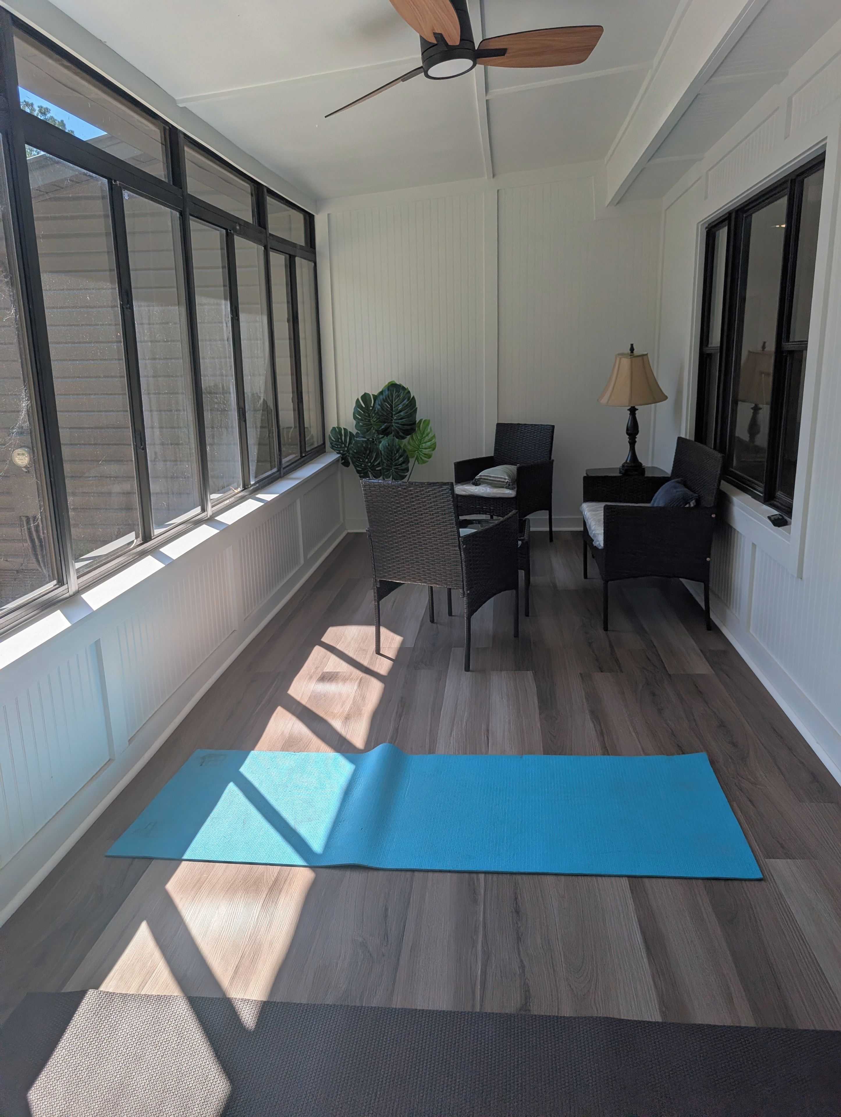 Una habitación iluminada por el sol con un ventanal grande, un ventilador de techo, una colchoneta de ejercicios azul y dos sillones de mimbre.