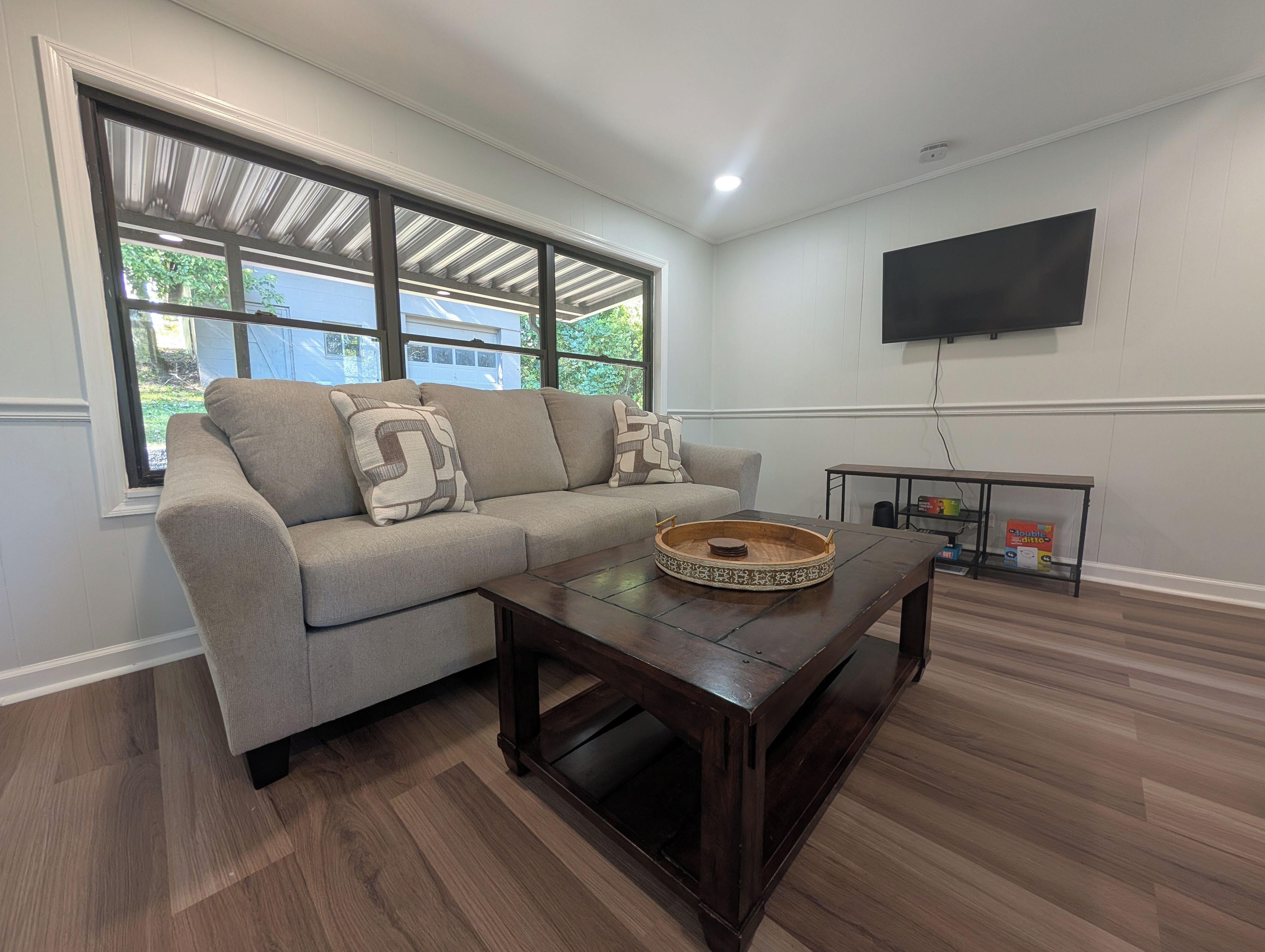 A living room with a sofa, a coffee table, a TV mounted on the wall, and a window with a view of greenery.