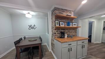 A dining area with a wooden table and chairs, a kitchen with a brick accent wall, and a shelf with framed pictures and a coffee maker.