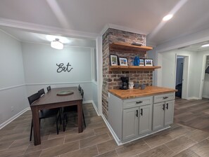 Un comedor con mesa y sillas de madera, una cocina con pared de ladrillo decorativa, y una estantería con marcos y una cafetera.