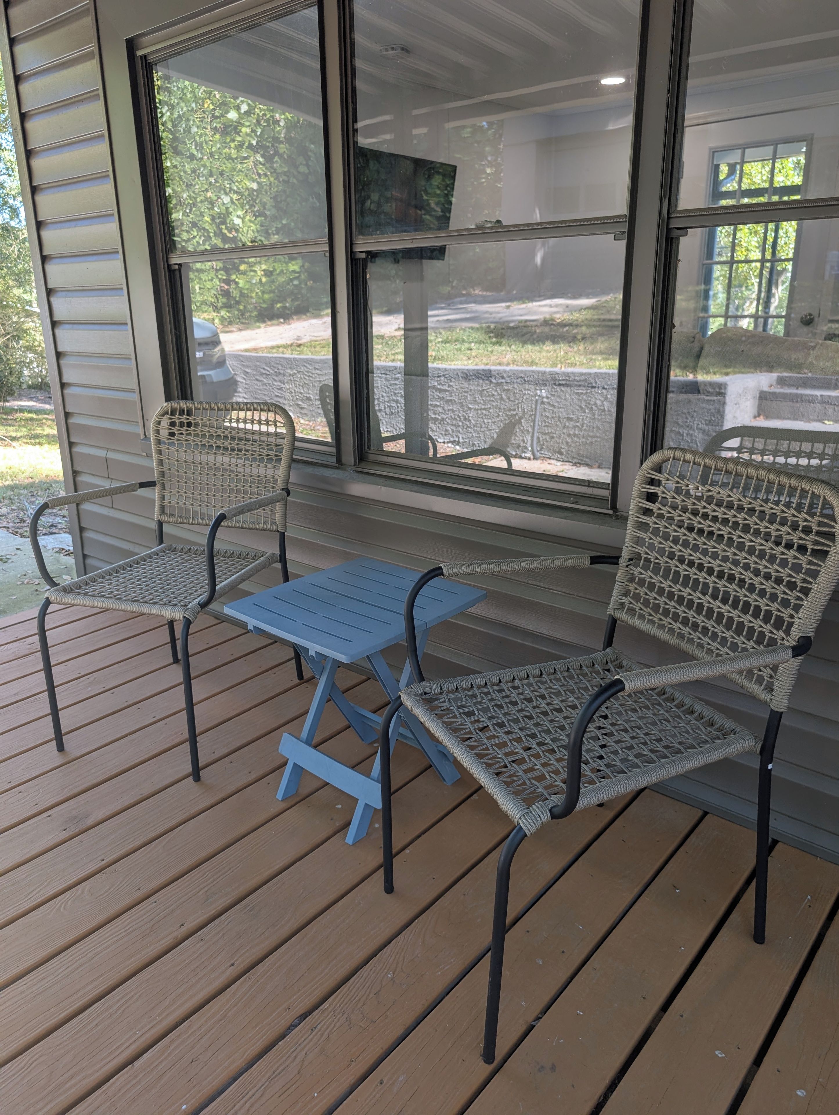 A covered outdoor seating area with two wicker chairs and a small table, overlooking a parking area.