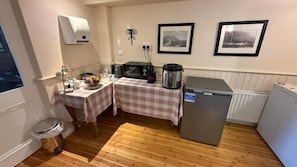 Cocina compacta con microondas, tostadora y hervidor sobre una mesa, un refrigerador y un cubo de basura.