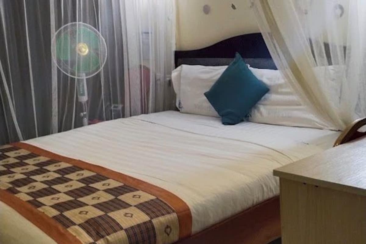 Een slaapkamer met een bed, nachtkastje, ventilator en gordijnen.