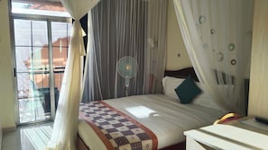Een slaapkamer met een bed, nachtkastje, ventilator en een raam met gordijnen.