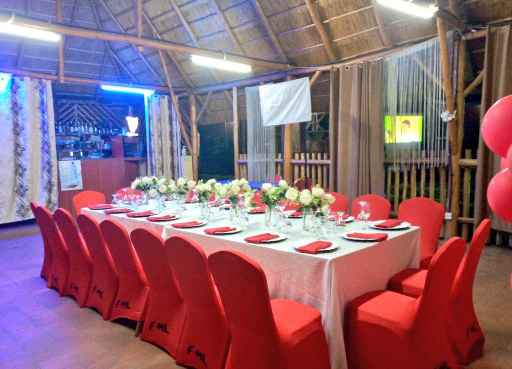 Un salón de banquetes con una larga mesa dispuesta para un evento formal, rodeado de sillas rojas, y una zona de bar visible al fondo.