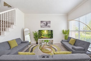 Ein modernes Wohnzimmer mit einem großen Flachbildfernseher, grauen Sofas, einem Spiegel und einer Treppe.
