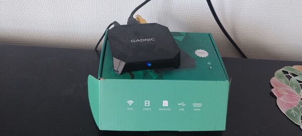 En sort elektronisk enhed med et blåt lys på en turkis kasse med ikoner for WIFI, VIDEO, MICROSD, USB og HDMI.