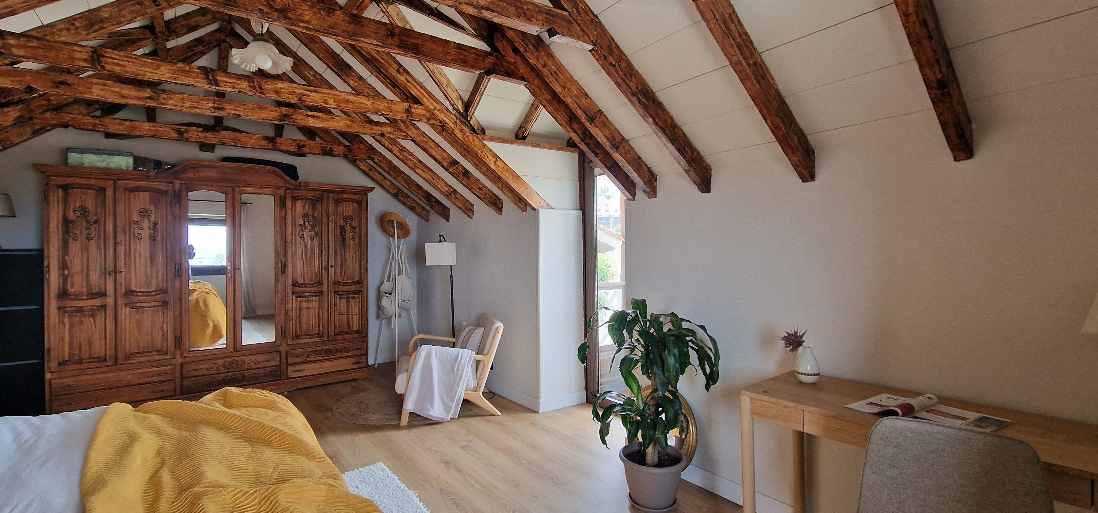 A room with exposed wooden beams, a wooden wardrobe, a bed with a yellow blanket, a potted plant, and a desk with a lamp.
