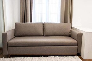 A beige sofa with a wooden base, positioned in front of a window with curtains. - Urban Luxury Apartments (Graz)
