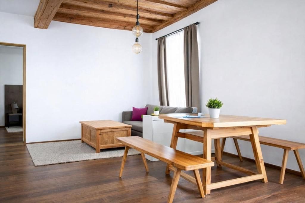 Espaço de estar moderno com mesa de jantar de madeira, bancos, sofá e pequena mesa lateral com uma planta.