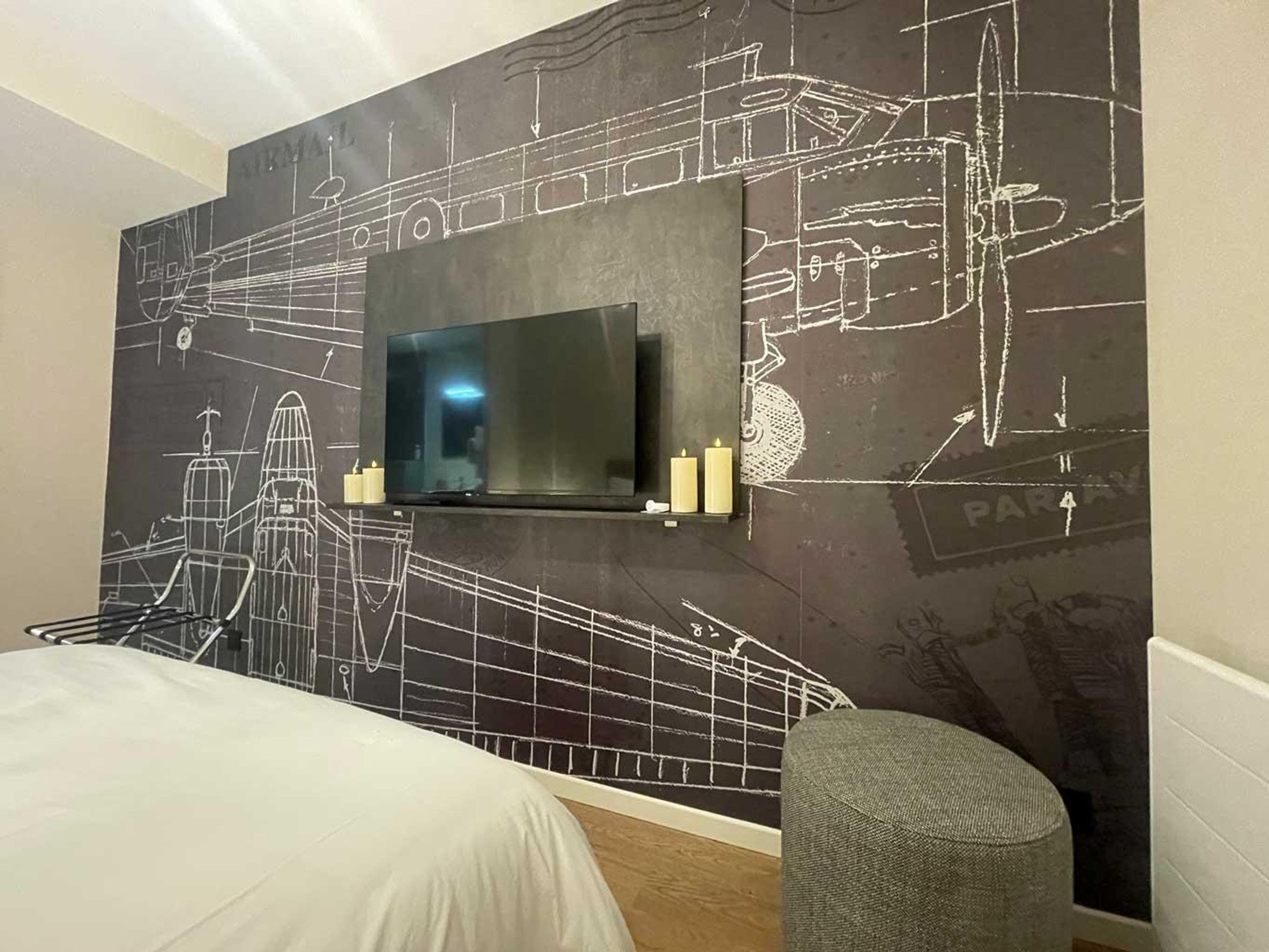 Een hotelkamer met een groot bed, een aan de muur bevestigde televisie met een stadsgezichtsmural, een klein zitgedeelte en een nachtkastje met kaarsen.