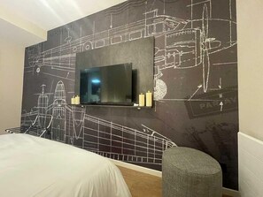 Una habitación de hotel con una cama grande, un televisor montado en la pared con un mural de la ciudad, un pequeño rincón de descanso y una mesita de noche con velas.