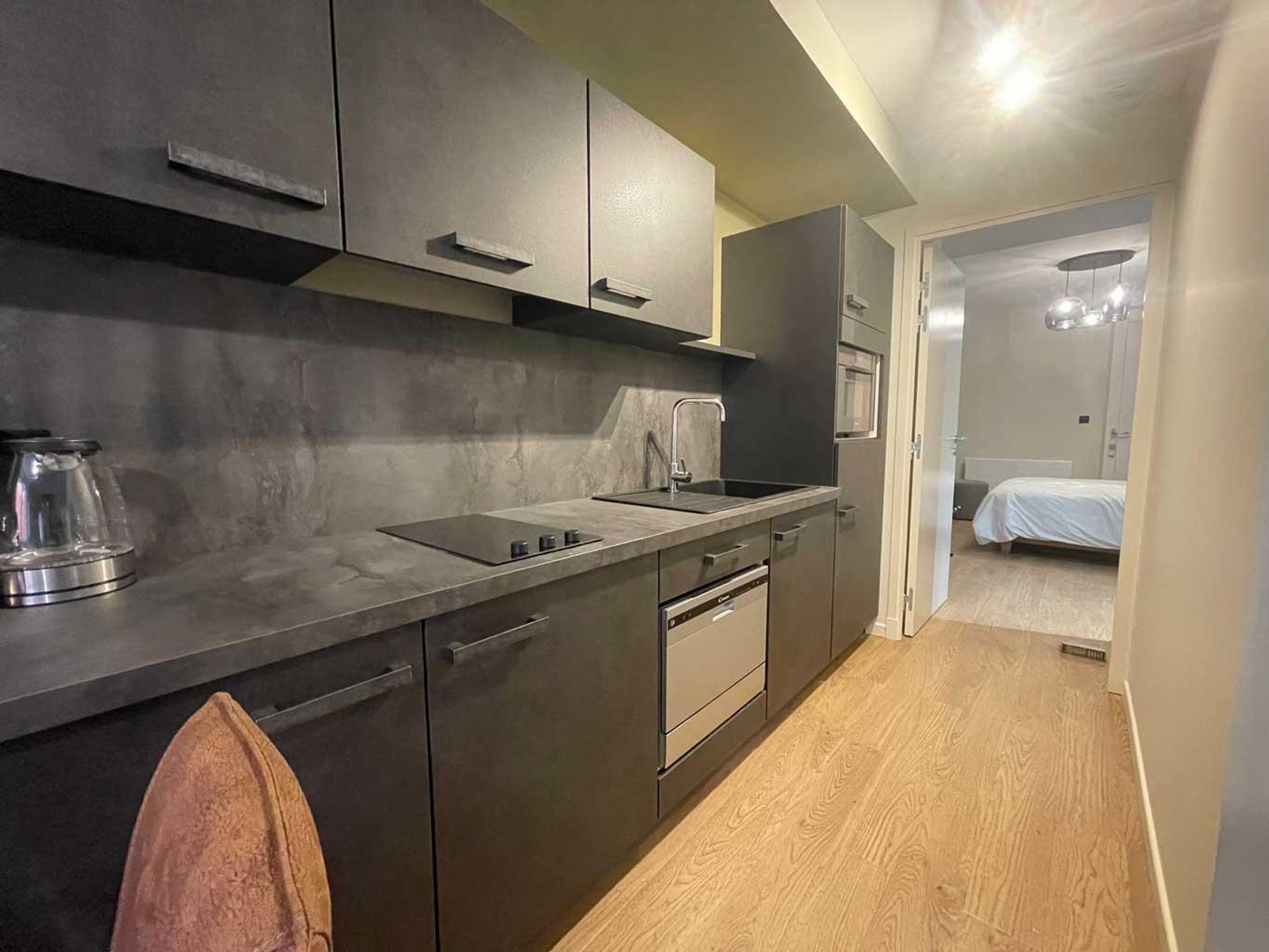 Une cuisine compacte équipée d’un évier, d’une cuisinière et de placards, qui donne sur une chambre avec un lit et une table de chevet.
