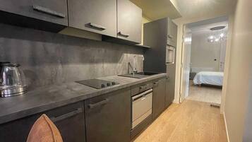 A compact kitchen with a sink, stove, and cabinets, leading to a bedroom with a bed and a nightstand.