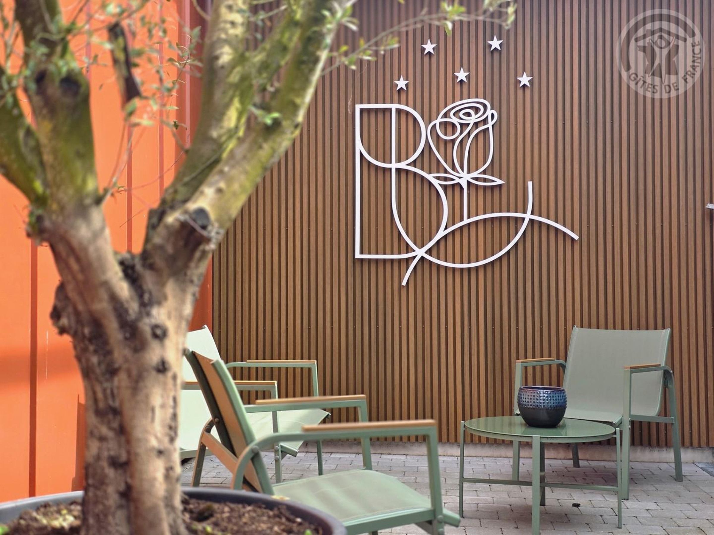 Un mur en bois orné d’un logo, une plante en pot et un espace extérieur aménagé avec des chaises et une table.