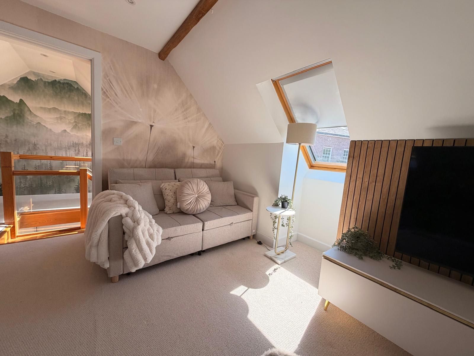 A modern loft bedroom with a sloped ceiling, a large window, a sofa, a TV, and a decorative wall.