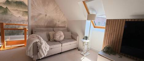 A modern loft bedroom with a sloped ceiling, a large window, a sofa, a TV, and a decorative wall.