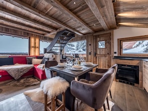 A cozy cabin interior with a dining area, a fireplace, and a large window overlooking a snowy landscape. - Cozy chalet mountain view - 5 guests (Courchevel)