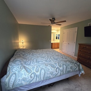 Ein Schlafzimmer mit einem Bett, Deckenventilator, Nachttisch, Kommode und einem Badezimmer im Hintergrund.