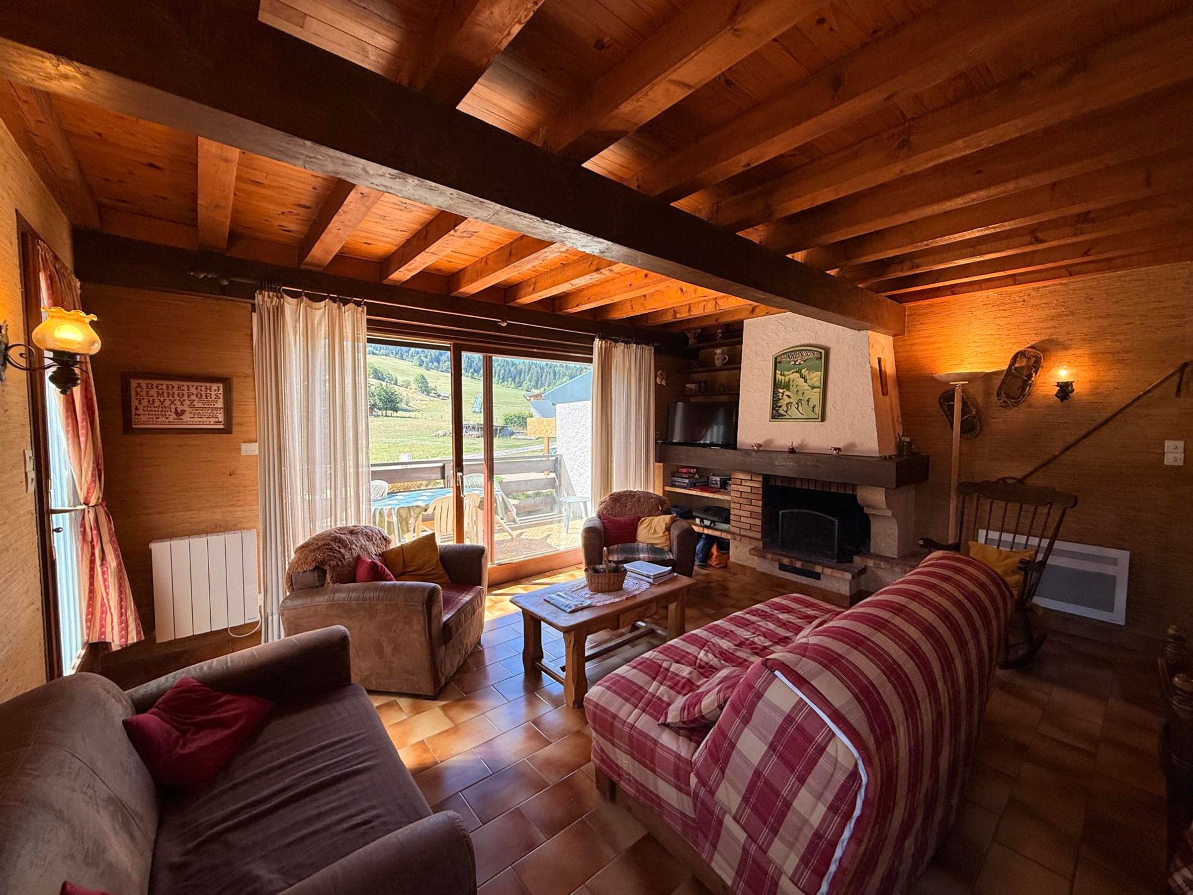 A cozy cabin interior with a fireplace, wooden beams, a sofa, a dining table, and a view of the outdoors.