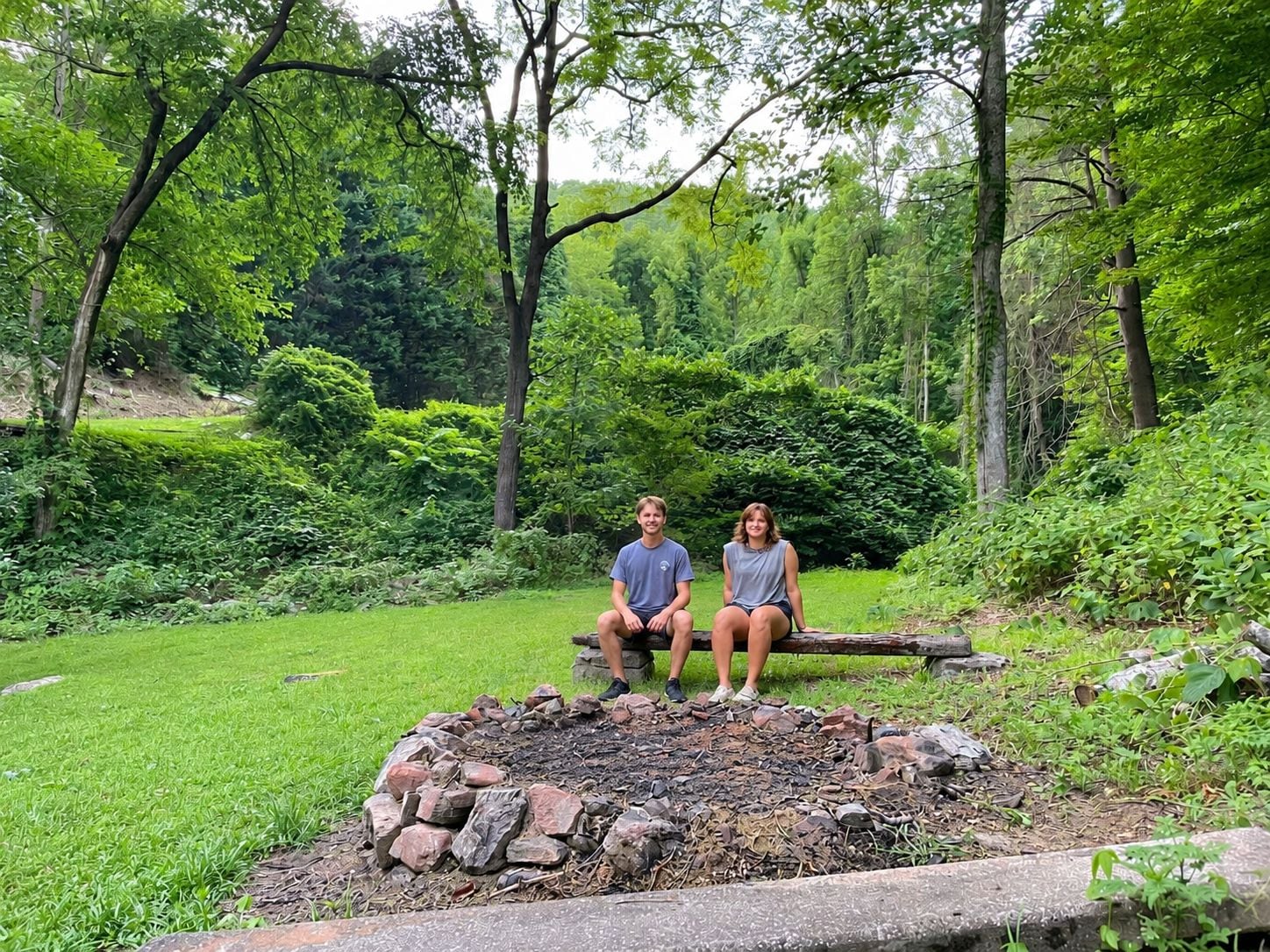 Two people sitting on a bench in a garden with a stone fire pit in the foreground.
