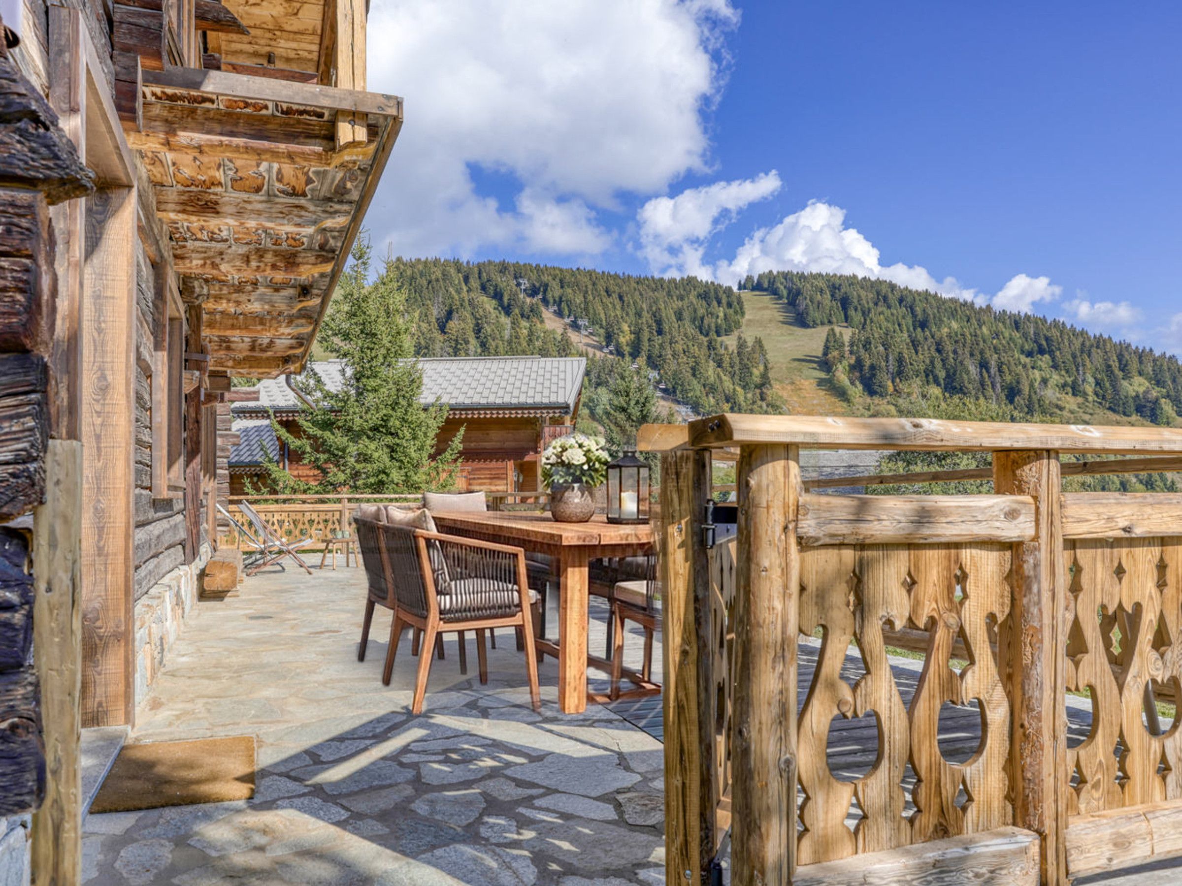 A wooden cabin with a stone patio, outdoor seating, and a view of a forested mountain.