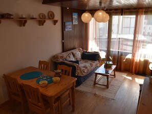 A cozy living room with a wooden dining table, chairs, a sofa, and a coffee table. There are decorative items on the wall and a window with curtains. - Central apartment - 6 guests (Montgenèvre)