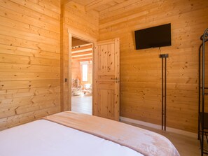 Ein Zimmer einer Holzhütte mit einem Bett, einem an der Wand befestigten Fernseher und einer Tür zu einem weiteren Raum.