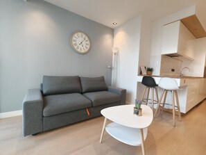 Ein moderner Wohnraum mit grauem Sofa, einem kleinen weißen Tisch und einer Küchenzeile mit weißer Arbeitsplatte und Barhocker.