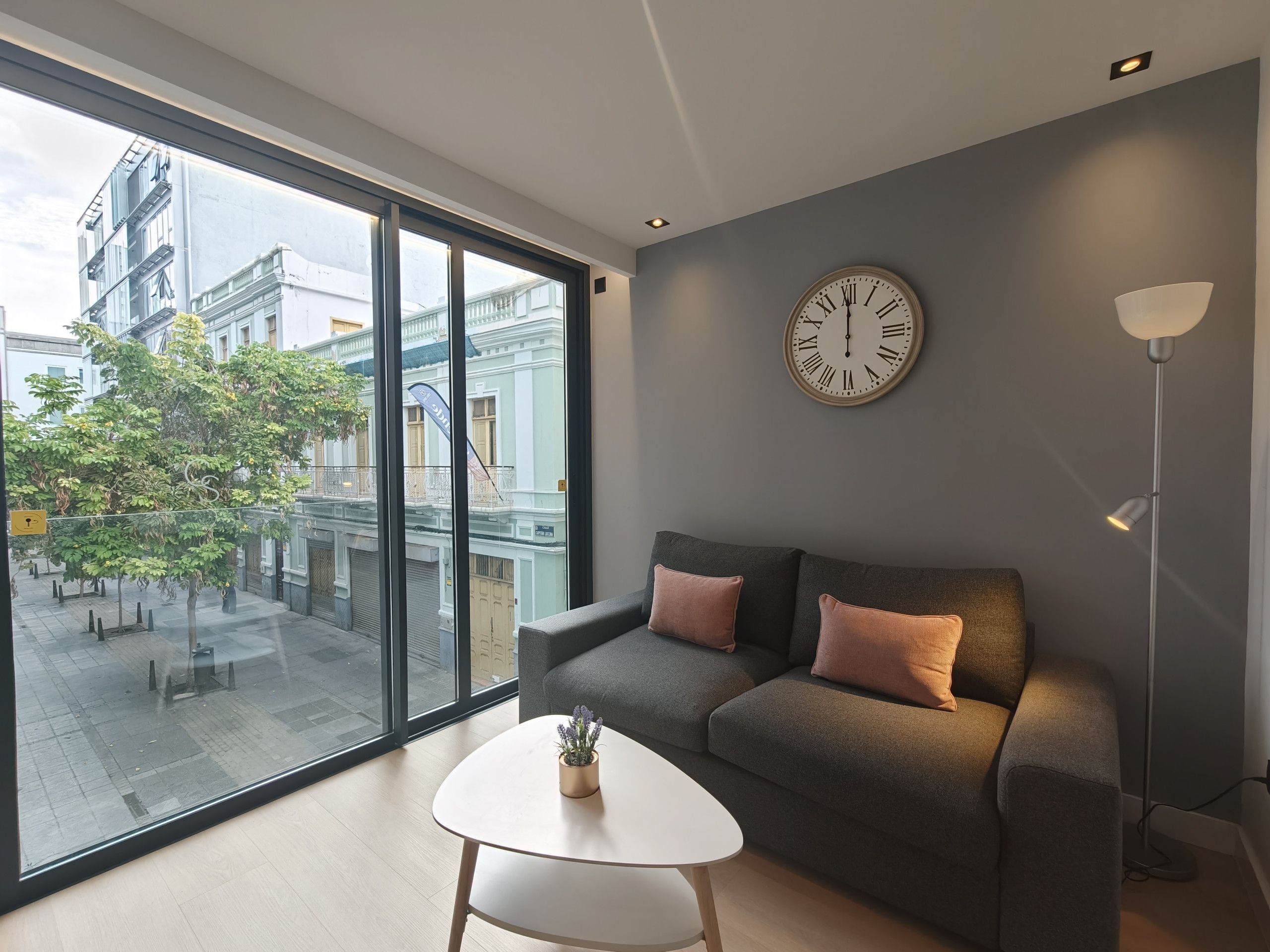 Un salon moderne avec un canapé gris, une petite table blanche et une grande fenêtre donnant sur des immeubles et des arbres.