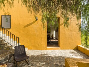 A stone-floored patio with a wicker chair, a staircase with a metal railing, and a doorway leading to an interior space with a chair and a table. - Casa África Loft - Finca Cruz Mayor (Telde)