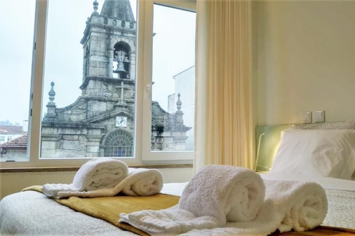 A hotel room with a bed, two rolled towels, a window with a view of a historic building, and a curtain.