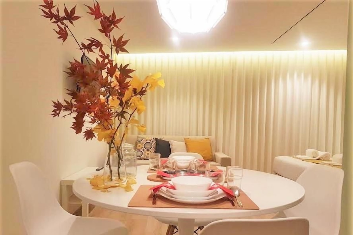 A dining area with a round table set for four, white chairs, a vase with autumn leaves, and a modern light fixture.