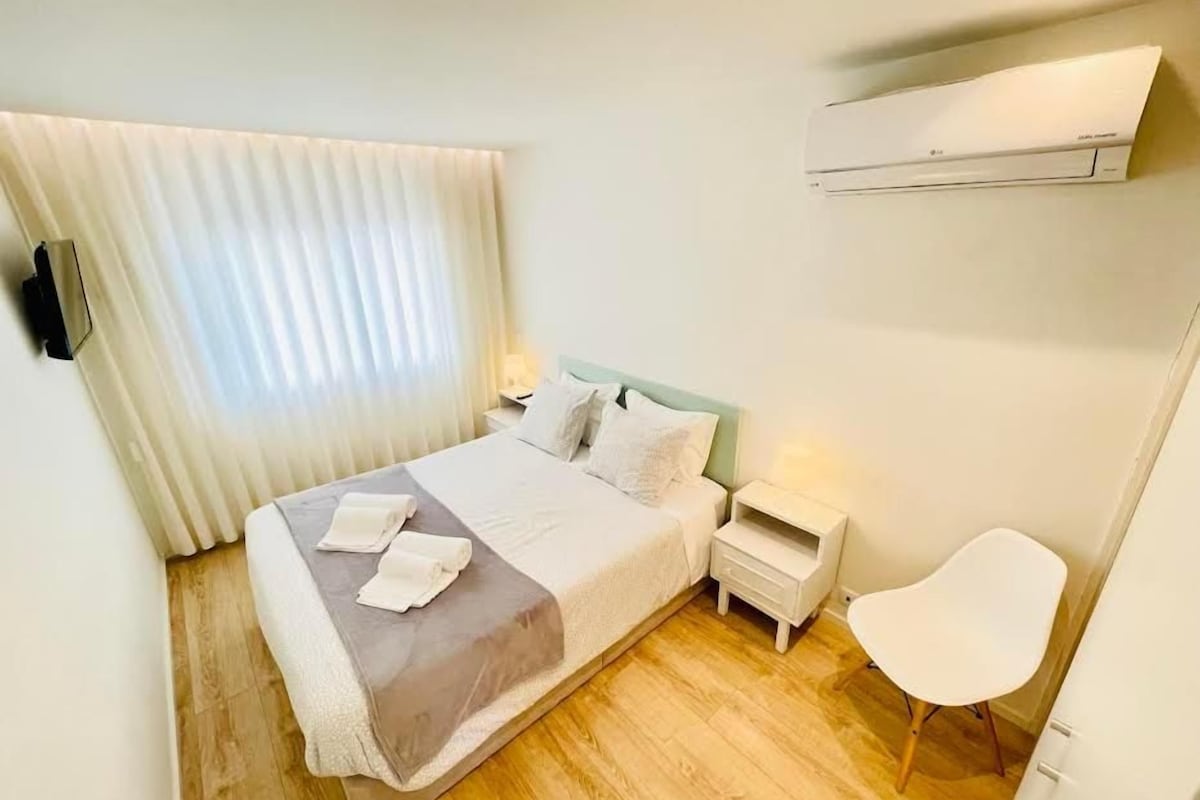 A hotel room with a bed, bedside tables, a chair, and an air conditioning unit.