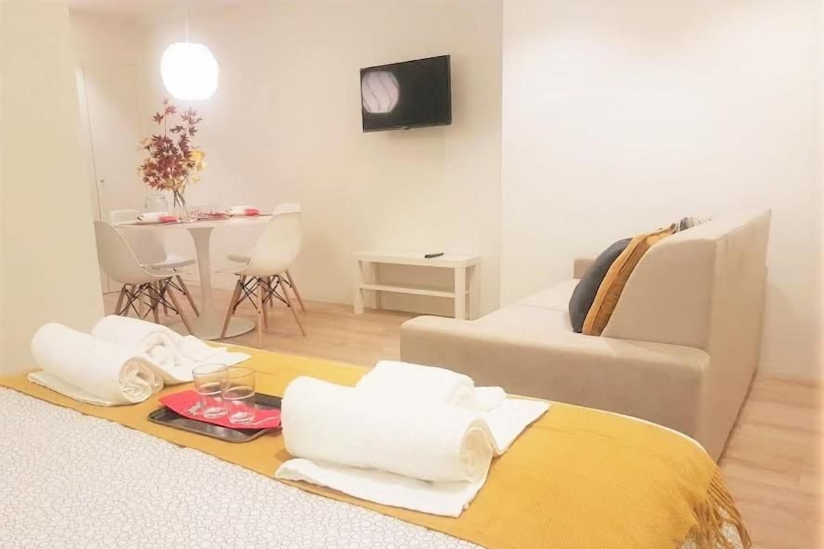 A hotel room with a bed, a sofa, a dining table with chairs, and a TV on the wall.